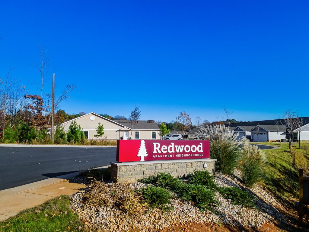 Red sign with white text reading "Redwood Apartment Neighborhoods" at the entrance of a residential area with houses and clear blue sky.