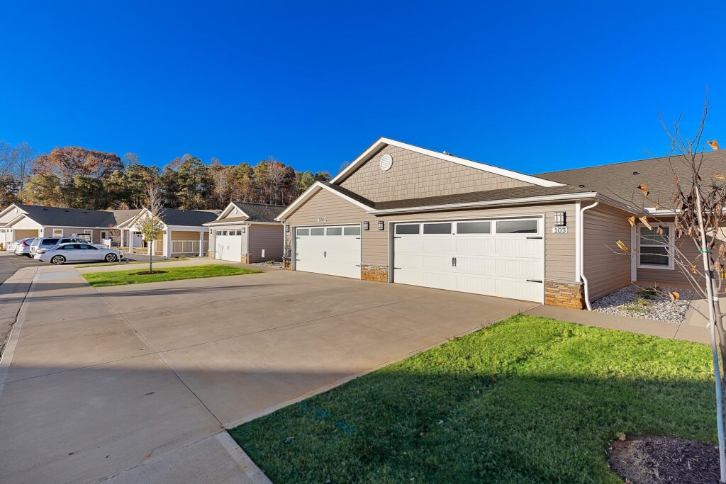 Single-story duplex homes with attached garages, concrete driveways, and manicured lawns under a clear blue sky.