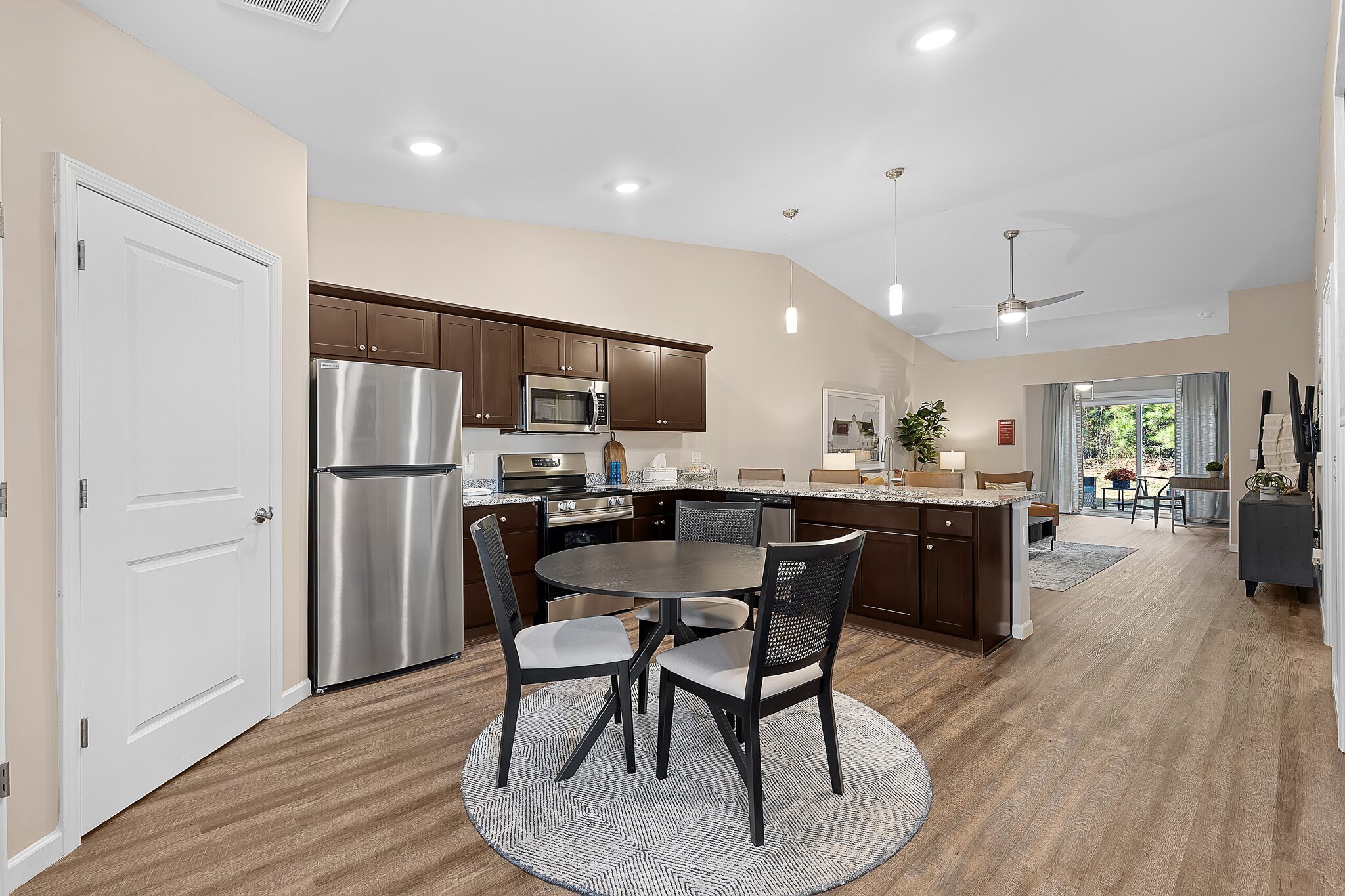 Modern kitchen and dining area with stainless steel appliances, dark wood cabinets, a round dining table with four chairs, and an open living space with large windows in the background.