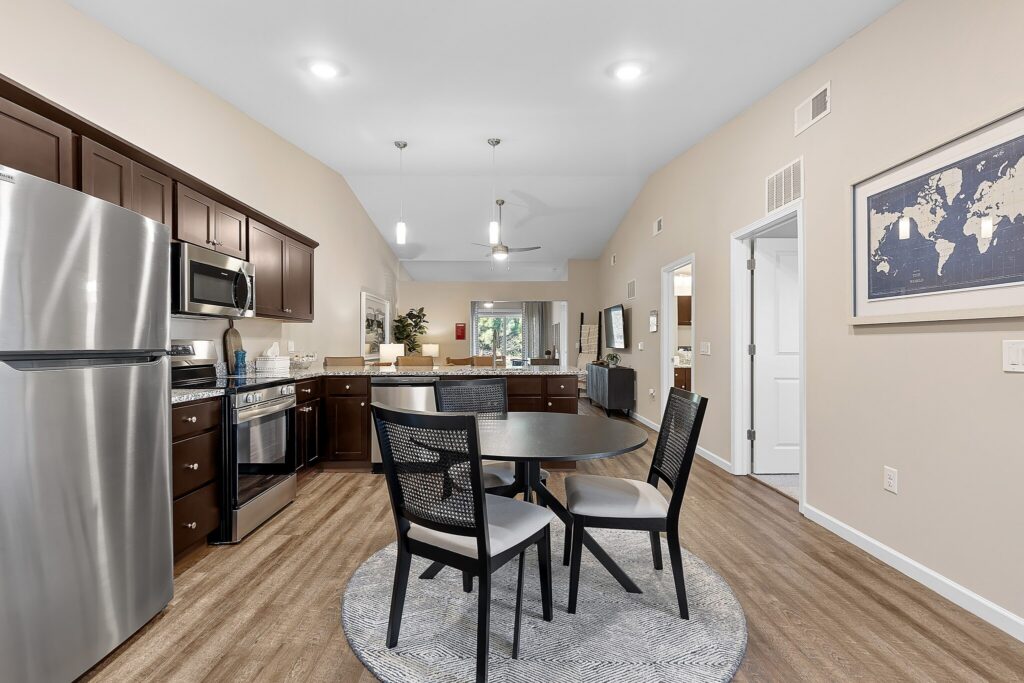 Modern kitchen and dining area with stainless steel appliances, dark wood cabinets, a round table with four chairs on a rug, and neutral-colored walls.