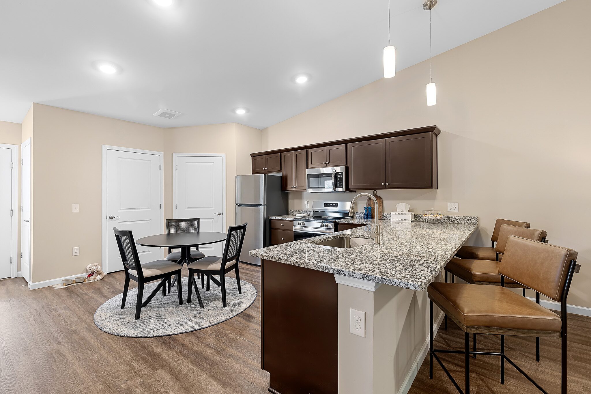 Modern kitchen and dining area with granite countertops, stainless steel appliances, round dining table with four chairs, and three bar stools on wooden flooring.