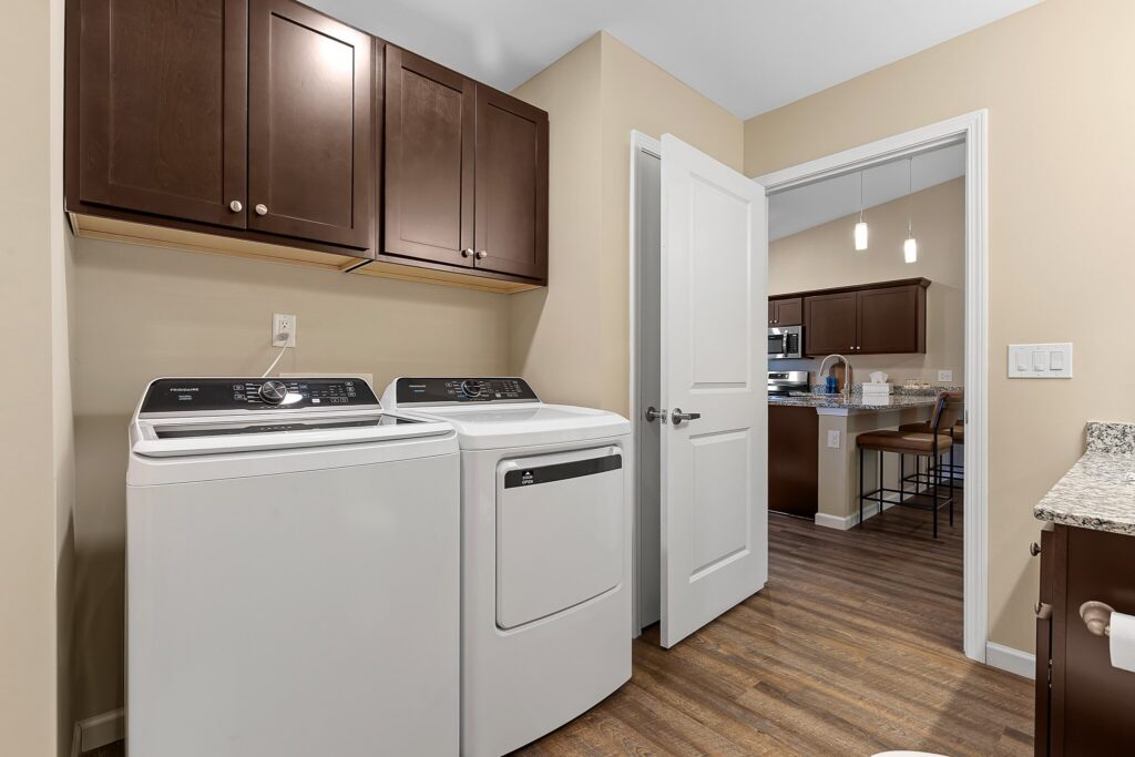 Laundry room with a washing machine and dryer, brown cabinets above, beige walls, and an open door leading to a kitchen area with wooden flooring.