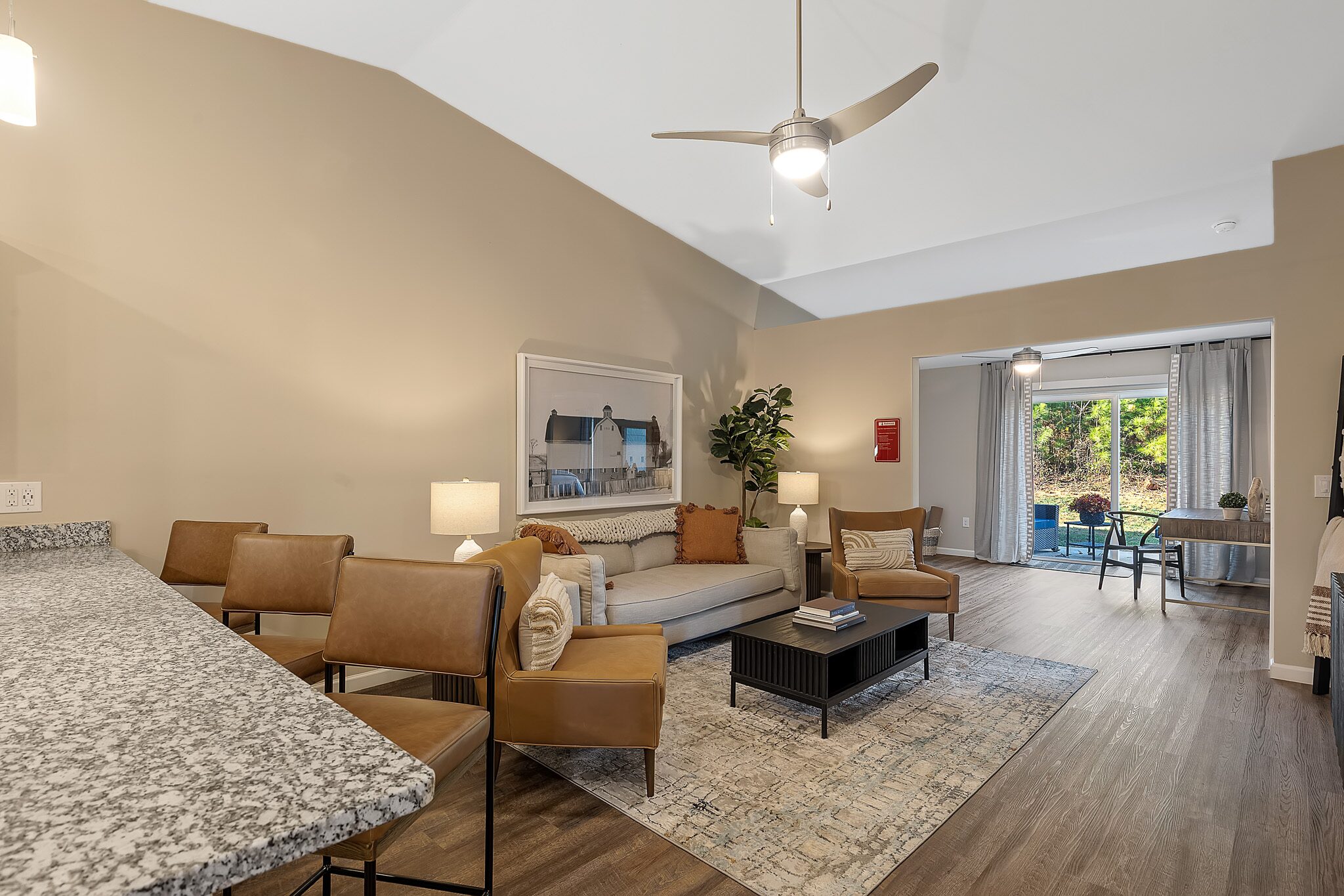 Modern living room with tan walls, brown leather chairs, a gray sofa, coffee table, area rug, and large windowed doors leading to a patio with outdoor seating and greenery.