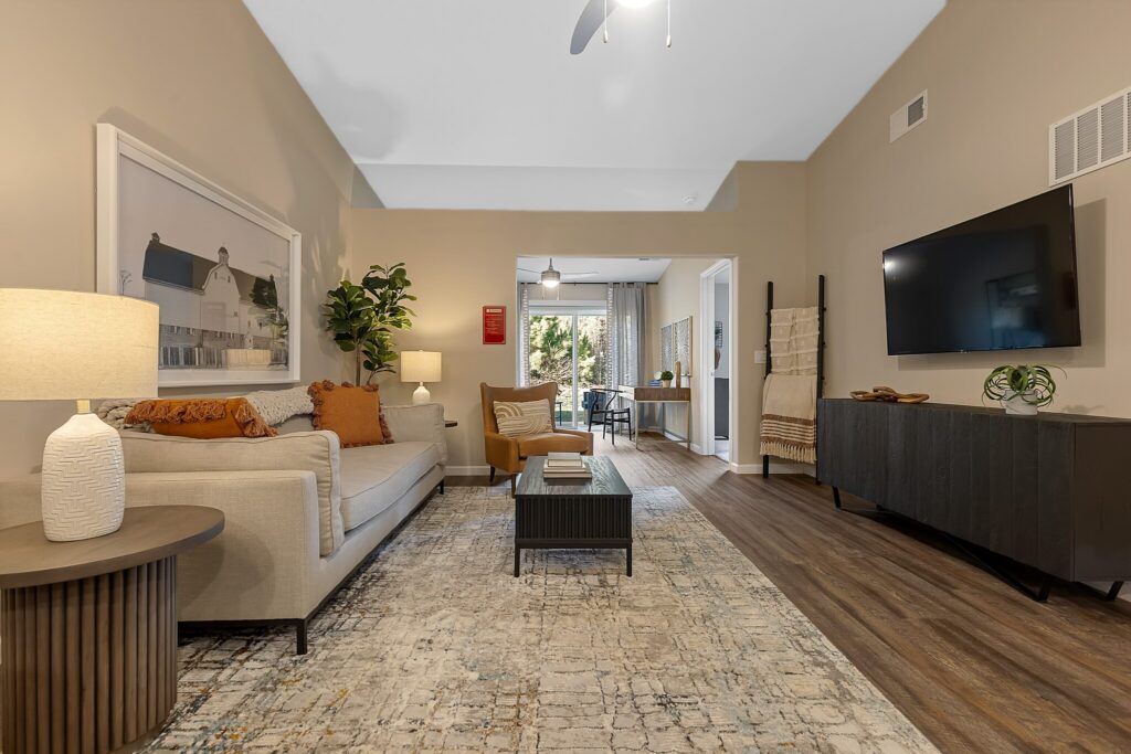 Neutral-toned living room with a sofa, cushions, side table with lamp, wall art, a TV mounted on the wall, and a coffee table on a patterned rug. Dining area visible in the background.