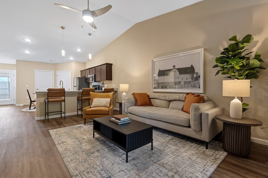 A modern living room with a gray sofa, armchair, coffee table, and floor plant; open kitchen with bar stools and pendant lights in the background.