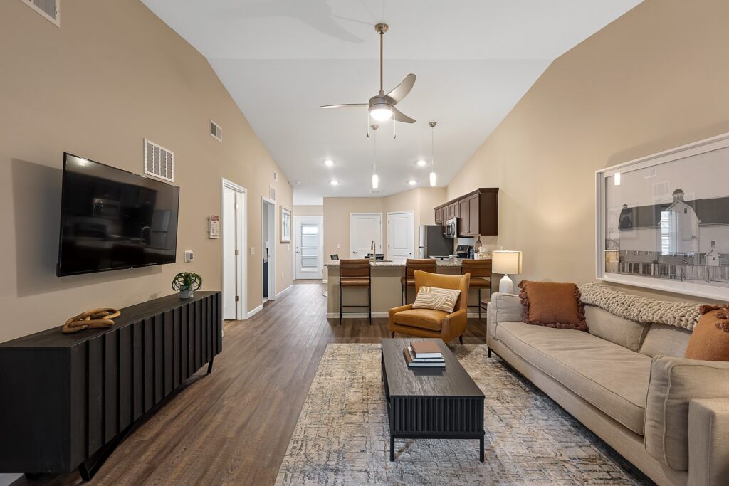 A modern living room with a beige sofa, black coffee table, wall-mounted TV, and open kitchen area with barstools; wood flooring and neutral-colored walls.