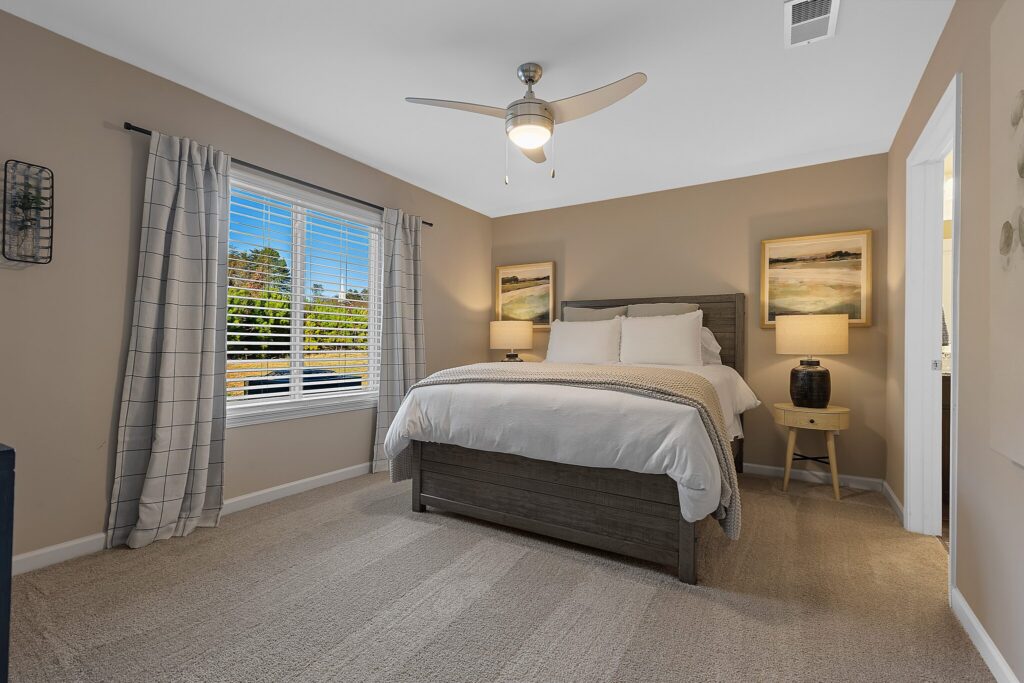 A bedroom with a large bed, two bedside tables with lamps, a ceiling fan, carpeted floor, large window with curtains, and two landscape paintings above the bed.