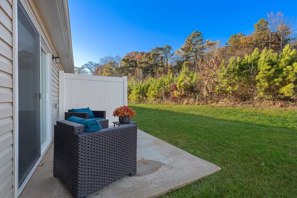 A small patio with two wicker chairs, a side table, and a white privacy panel overlooks a grassy yard bordered by trees under a clear blue sky.