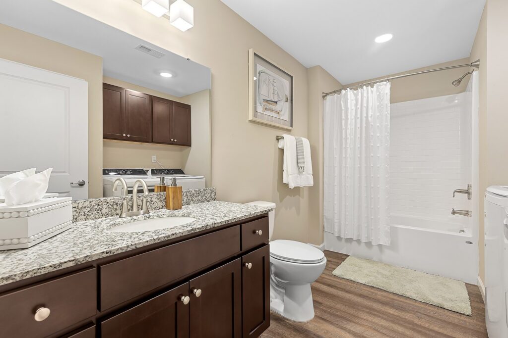 Modern bathroom with granite countertop, dark wood cabinetry, toilet, bathtub with white shower curtain, and stacked washer and dryer reflected in the large mirror.
