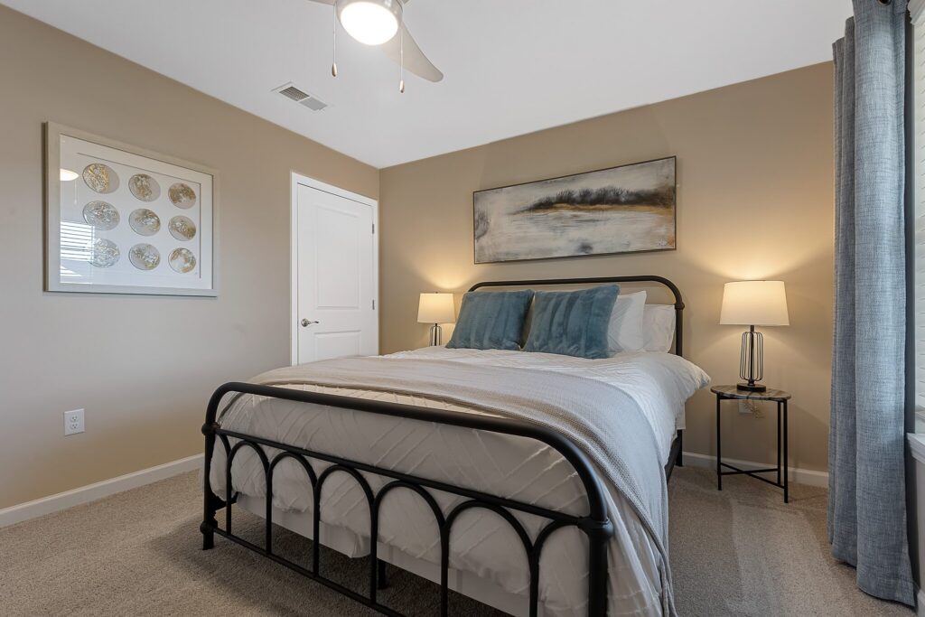 A neatly made bed with a metal frame, two blue accent pillows, two bedside tables with lamps, beige walls, framed art, and a window with gray curtains.