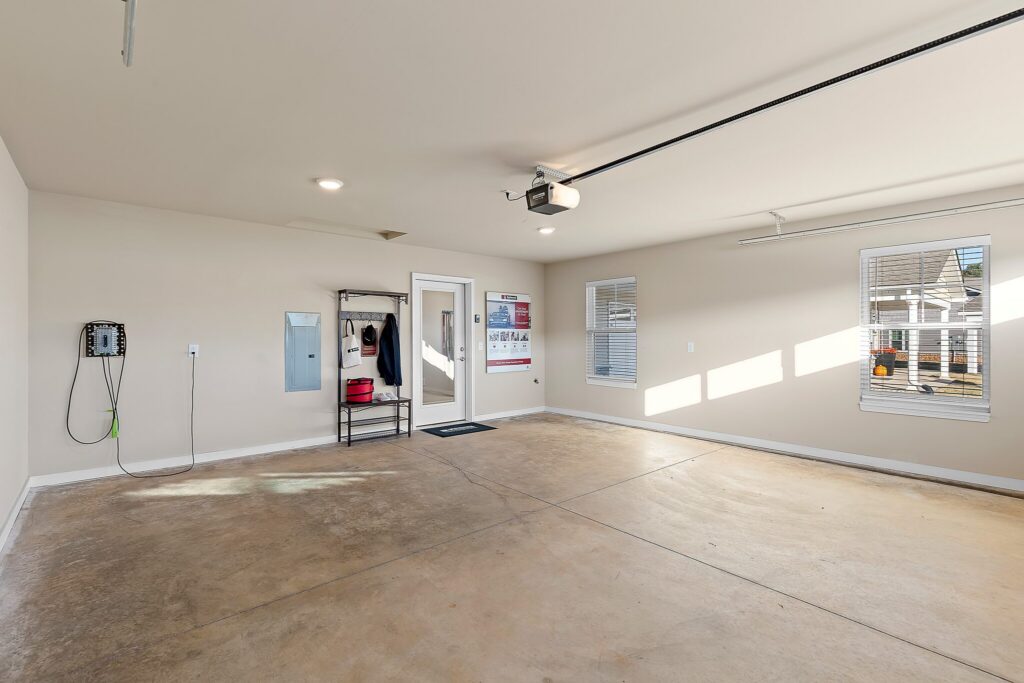 Clean, empty garage with beige walls, concrete floor, coat rack, wall-mounted charger, two windows, overhead garage door opener, and a door leading inside.