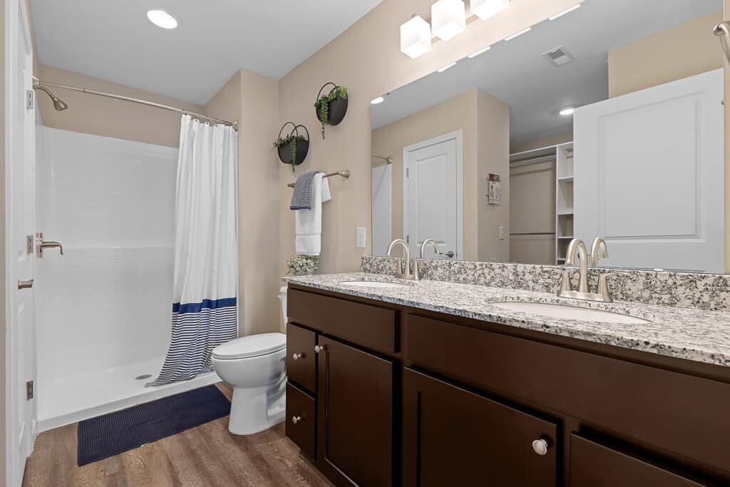 Modern bathroom with double sink vanity, granite countertop, toilet, walk-in shower with curtain, two wall-mounted plant holders, and wood-look flooring.