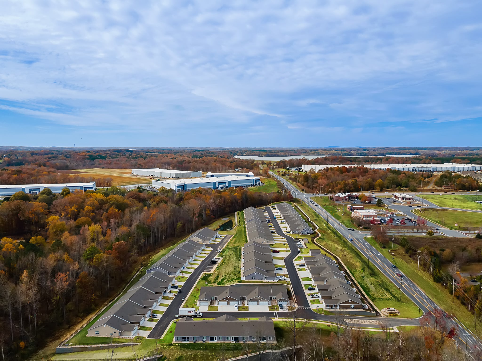 Aerial view of a suburban neighborhood with rows of houses, nearby commercial buildings, a main road, and surrounding autumn trees.