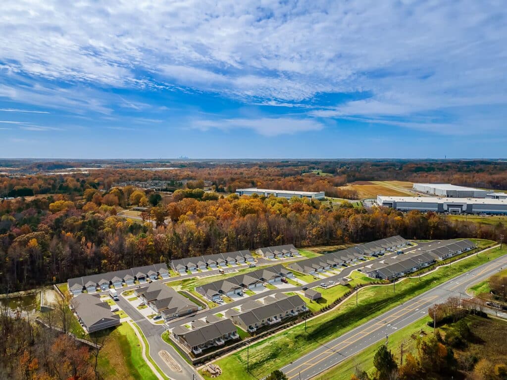 Aerial view of a suburban neighborhood with rows of houses, parking lots, and a main road, surrounded by trees with autumn foliage under a partly cloudy sky.