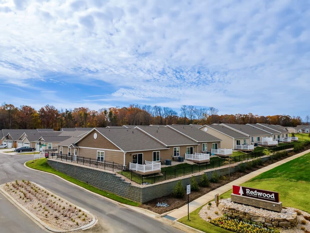 A row of single-story tan townhomes with fenced patios sits beside a road under a partly cloudy sky. A large "Redwood" sign is displayed in landscaped greenery at the entrance.