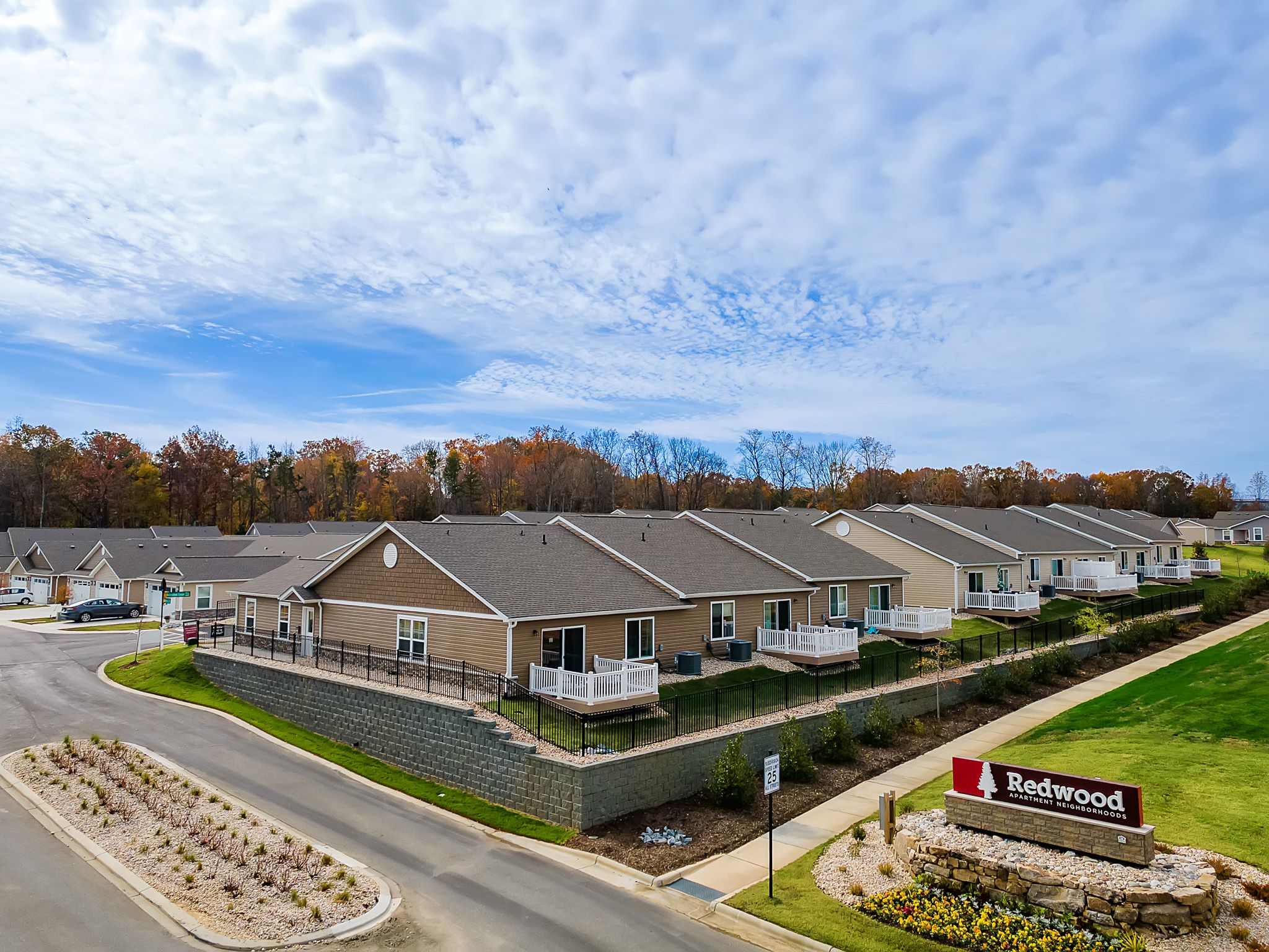 A row of single-story tan townhomes with fenced patios sits beside a road under a partly cloudy sky. A large "Redwood" sign is displayed in landscaped greenery at the entrance.