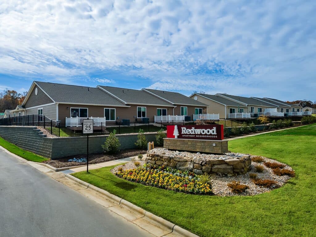Row of single-story homes behind a "Redwood Apartment Neighborhoods" sign, landscaped with flowers and stone, on a suburban street under a partly cloudy sky.