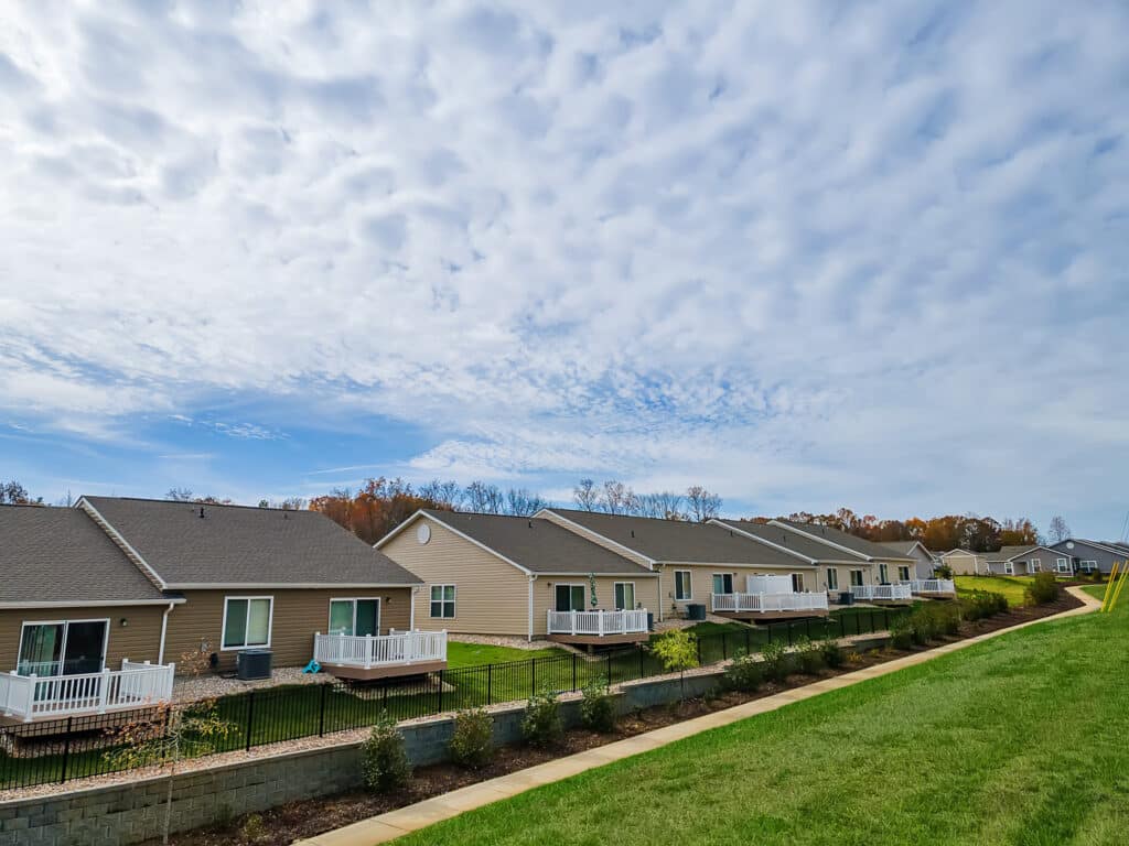 Row of single-story houses with fenced backyards, white decks, and a grassy area in the foreground under a partly cloudy sky.