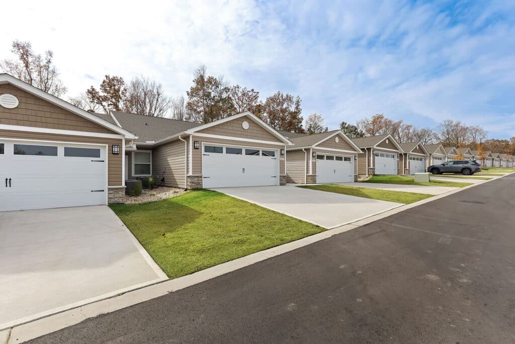 Row of single-story suburban houses with attached garages, neatly trimmed lawns, and a paved street under a partly cloudy sky.