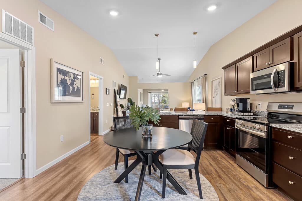 Modern kitchen and dining area with dark wood cabinets, round black dining table with four chairs, stainless steel appliances, and light wood flooring.