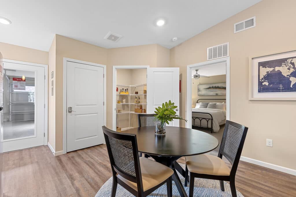 A round dining table with four chairs sits in an apartment next to a pantry, bedroom, and hallway. A plant centerpiece and wall art are visible.