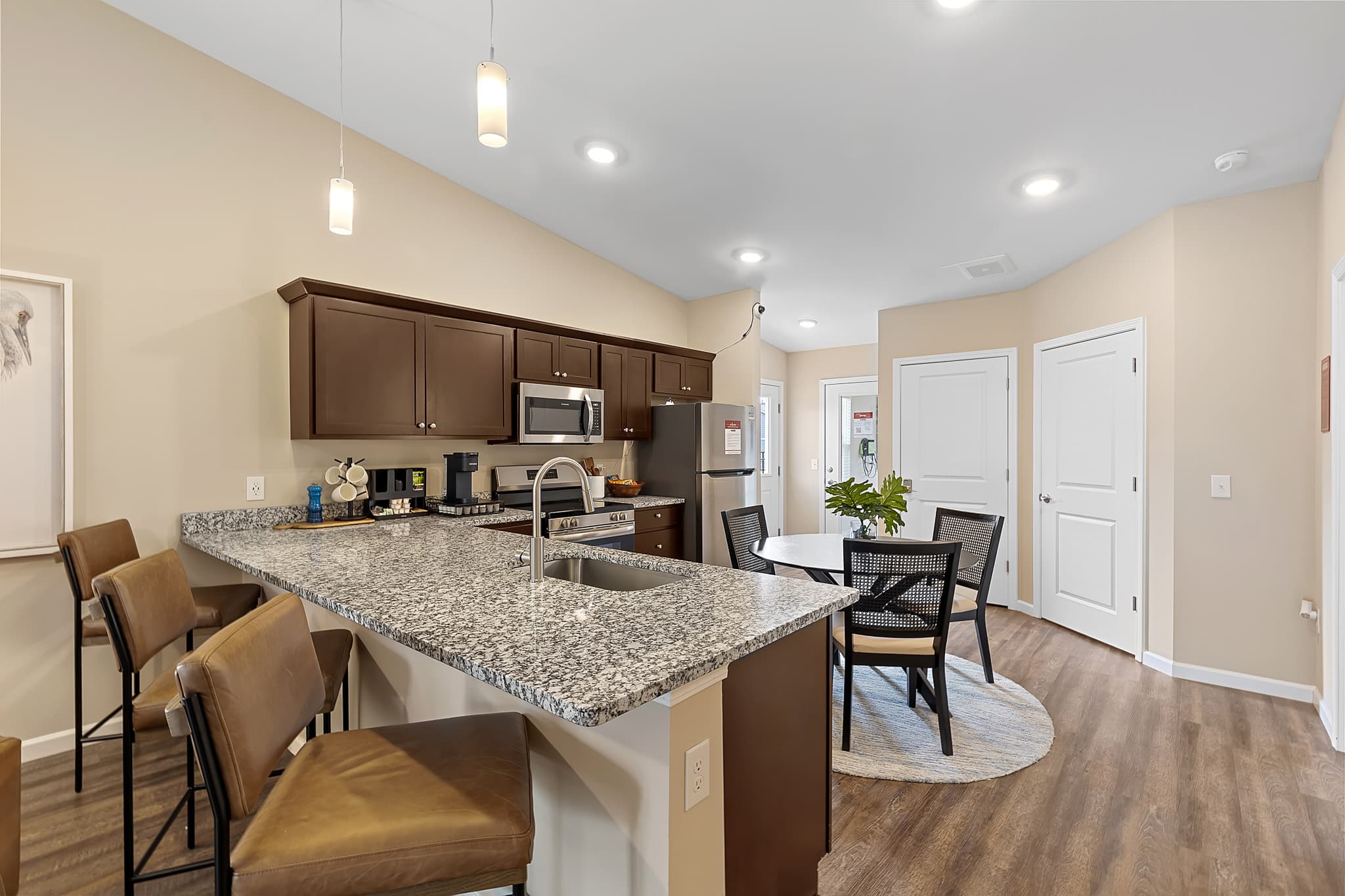 Modern kitchen with granite countertops, brown cabinets, stainless steel appliances, bar seating, and a round dining table with four chairs on a wooden floor.