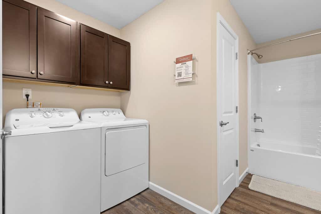 Laundry room with a white washer and dryer, brown cabinets above, wood flooring, and an adjacent bathroom with a white bathtub and beige walls.