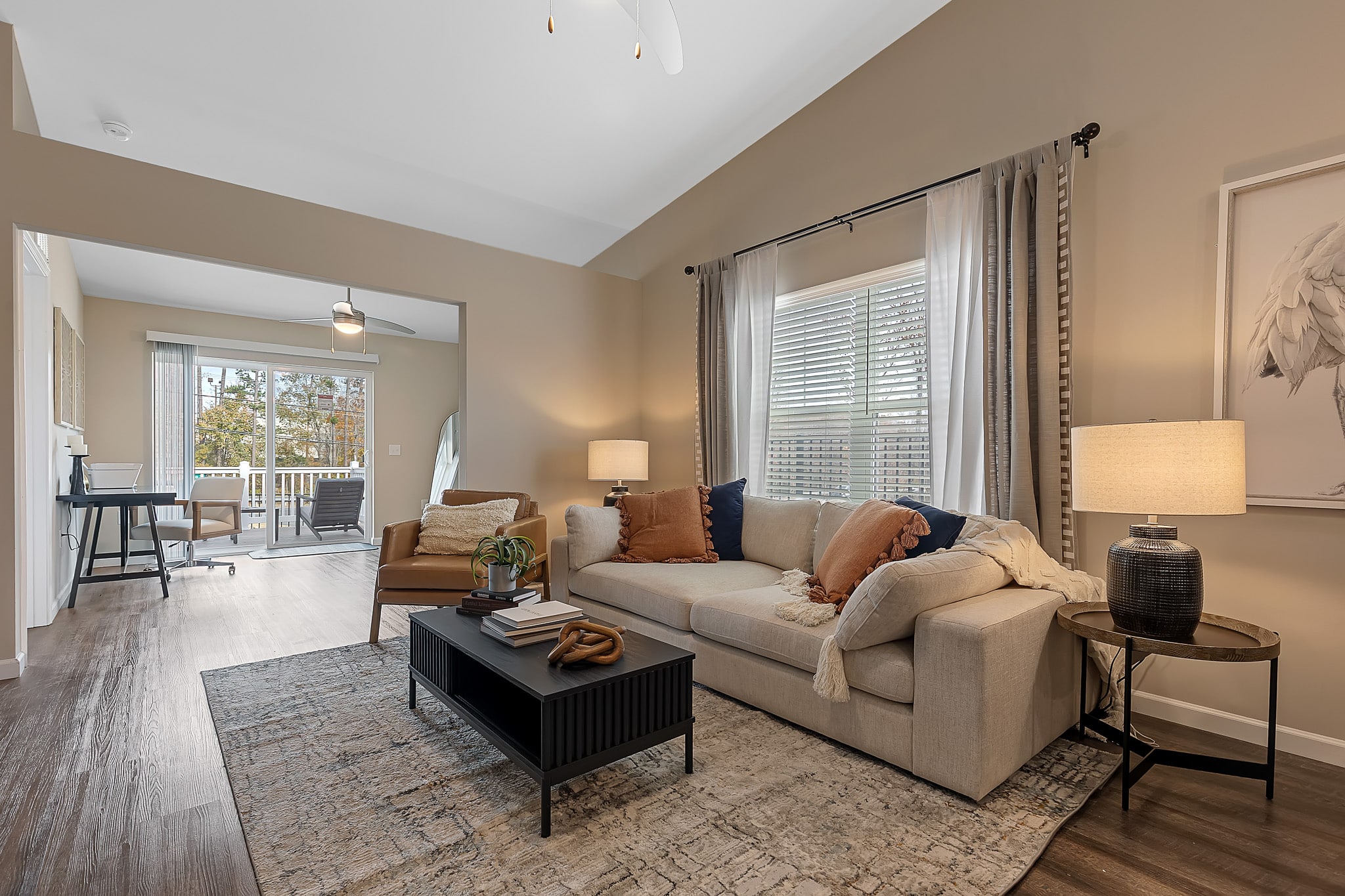 A modern living room with a beige sofa, a coffee table, a brown armchair, lamps, and curtains. An adjacent workspace and balcony are visible in the background.