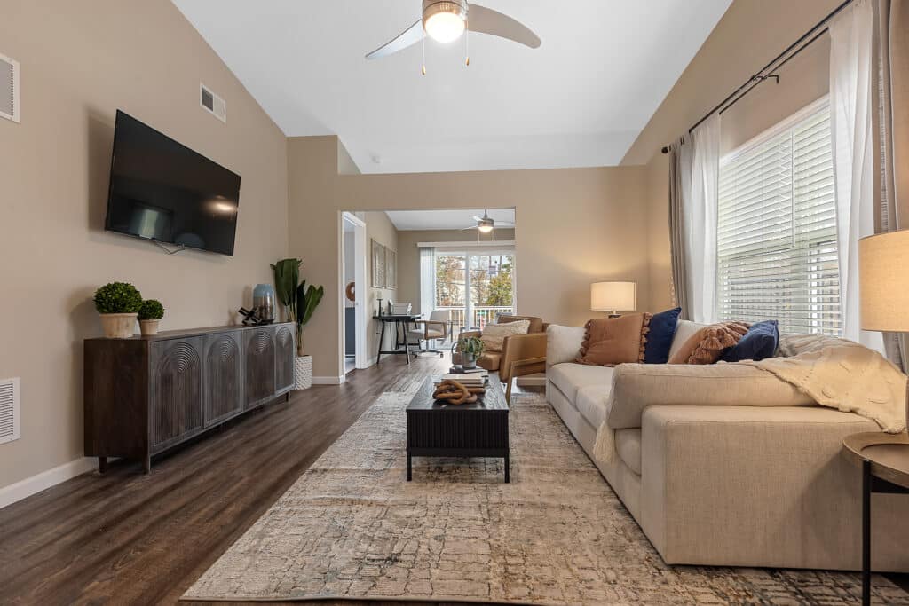A modern living room with a beige sofa, patterned rug, wall-mounted TV, wooden console, and large windows with sheer curtains, leading to a dining area in the background.