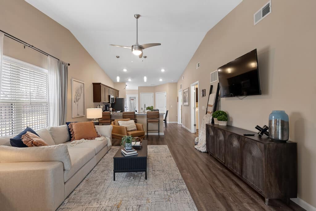Modern living room with beige walls, a mounted TV, light sofa, accent chair, and a rug. The space opens to a kitchen with wood cabinets and pendant lighting.