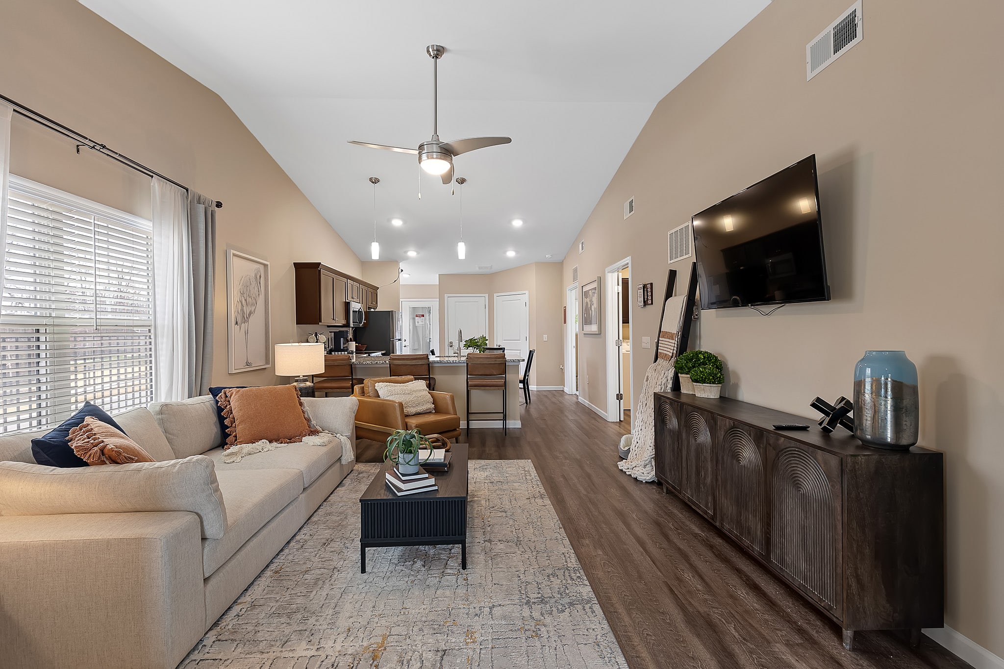 Modern living room with beige walls, a mounted TV, light sofa, accent chair, and a rug. The space opens to a kitchen with wood cabinets and pendant lighting.