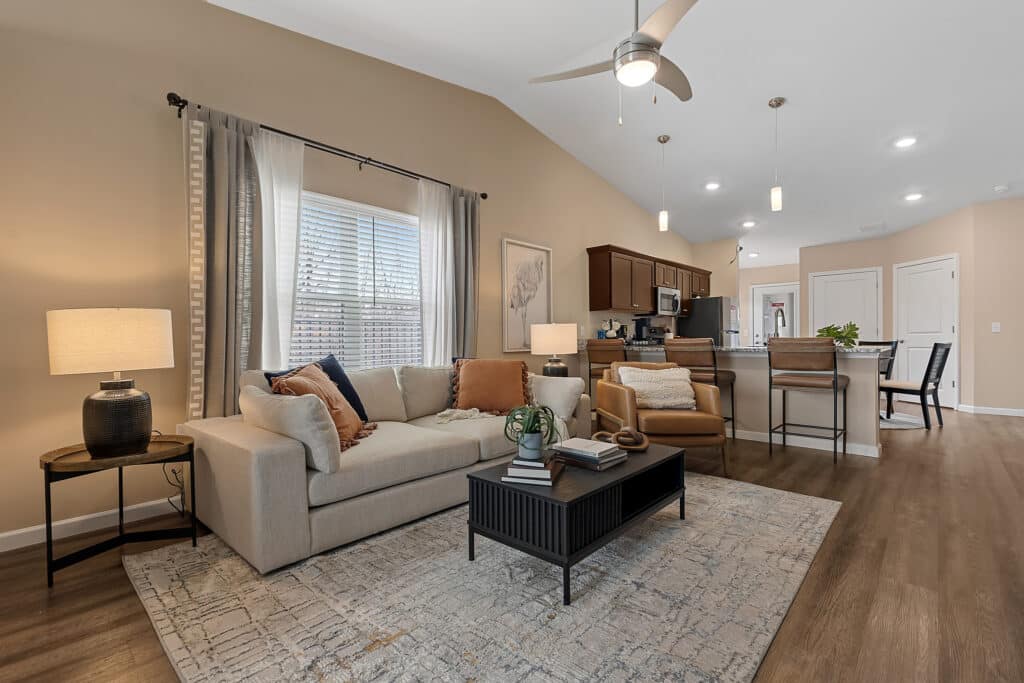 A modern living room with a beige sofa, coffee table, armchair, and side table, leading to an open kitchen with bar stools and pendant lights.