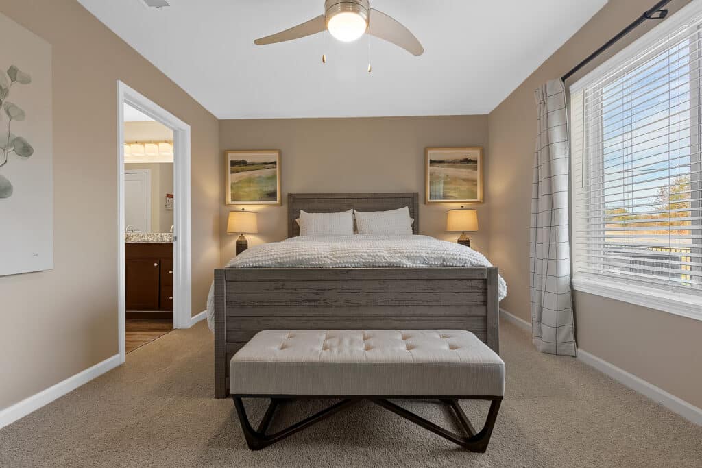 Neutral-toned bedroom with a wooden bed, white bedding, two bedside tables with lamps, a cushioned bench at the foot of the bed, and large window with white blinds.