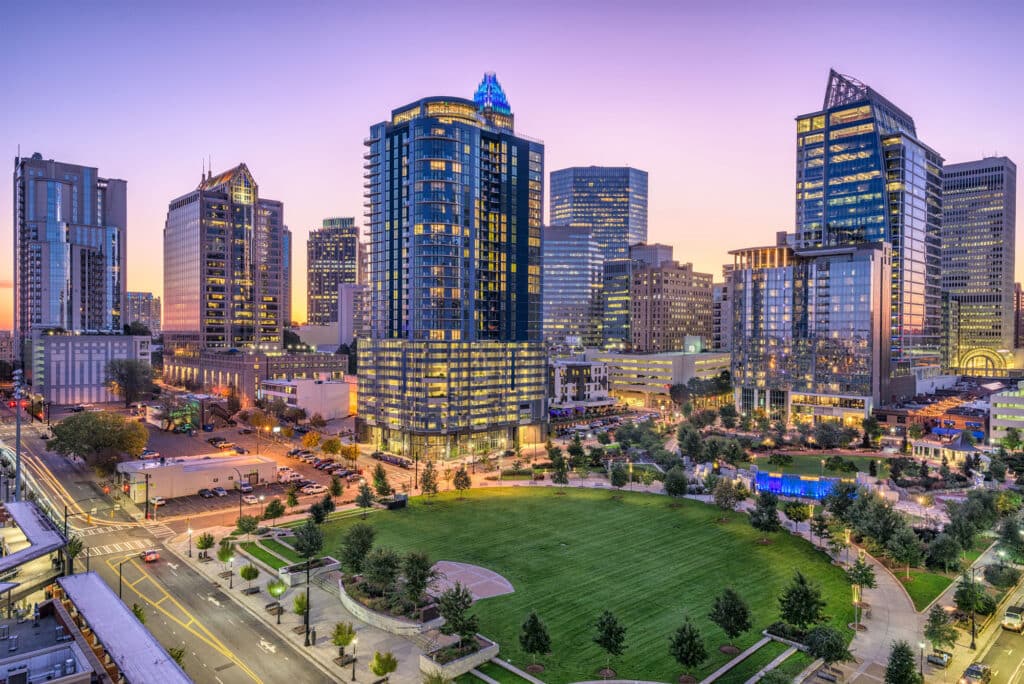 A cityscape at dusk features modern high-rise buildings surrounding a green park with walking paths and streetlights, under a purple and orange sky.