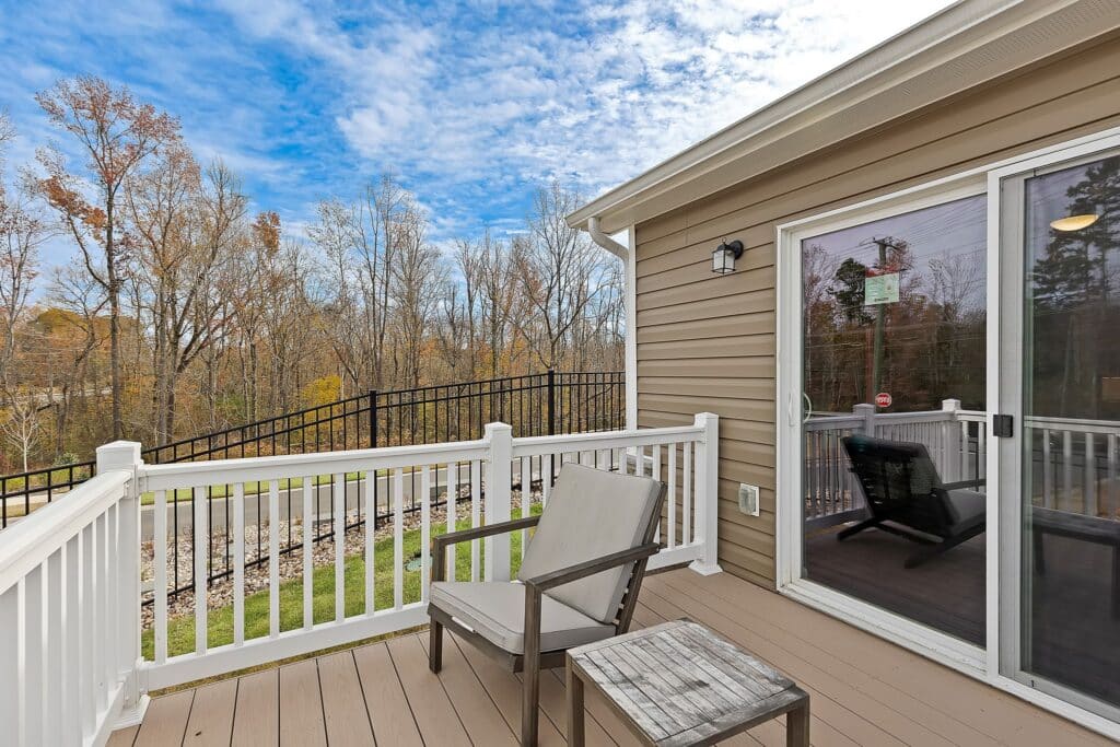 A small deck with two wooden chairs and a table, white railing, and sliding glass door, overlooking a yard with trees under a partly cloudy sky.