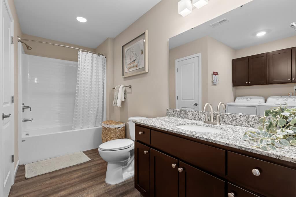 Modern bathroom with a granite countertop, double sink, dark wood cabinets, toilet, bathtub with white shower curtain, laundry area, and framed artwork above a towel rack.