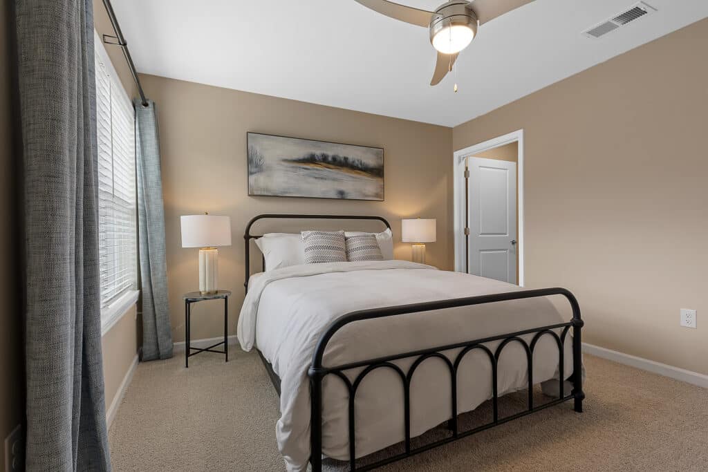 A neatly made bed with white bedding in a beige bedroom, flanked by two lamps on bedside tables, with a landscape painting above the headboard.