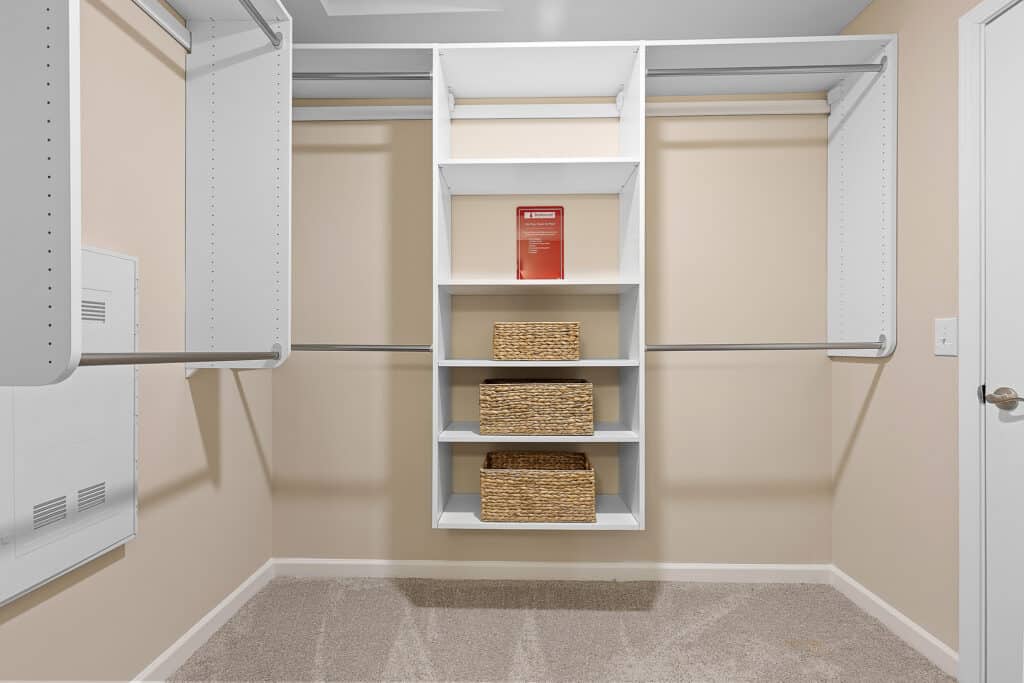 Empty walk-in closet with beige walls, carpeted floor, white shelving unit, three woven baskets, and a red item on the top shelf.