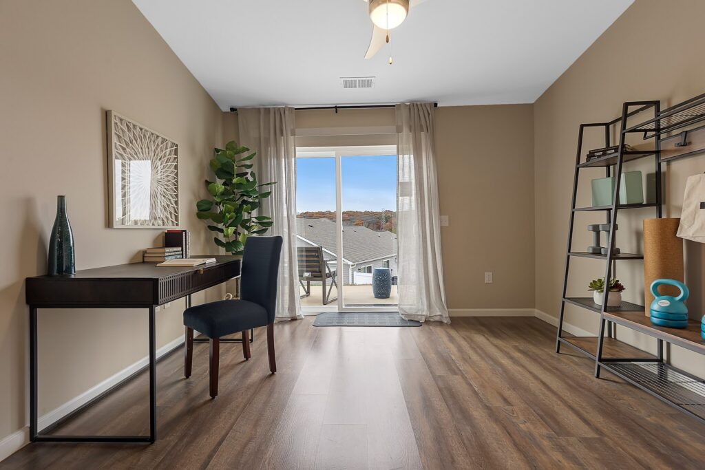 A home office with a desk, chair, plant, shelving unit, and sliding glass doors leading to a balcony with a view of rooftops and hills.