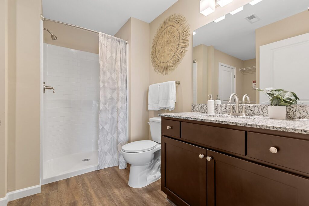 Modern bathroom with a walk-in shower, white toilet, dark wood vanity with granite countertop, large mirror, and decorative wall art above towel rack.