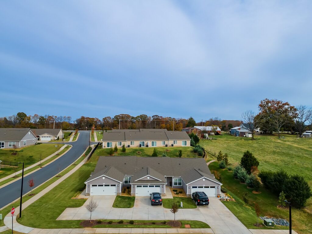 Aerial view of suburban homes with driveways and garages, bordered by a road and grassy areas, with trees and additional houses in the background.