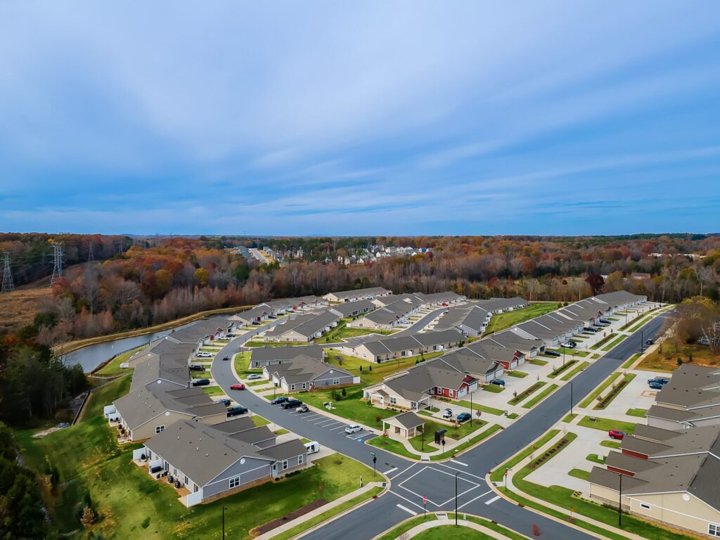 Aerial view of a suburban neighborhood with single-story houses, neatly arranged streets, green lawns, and a wooded area in the background under a cloudy sky.