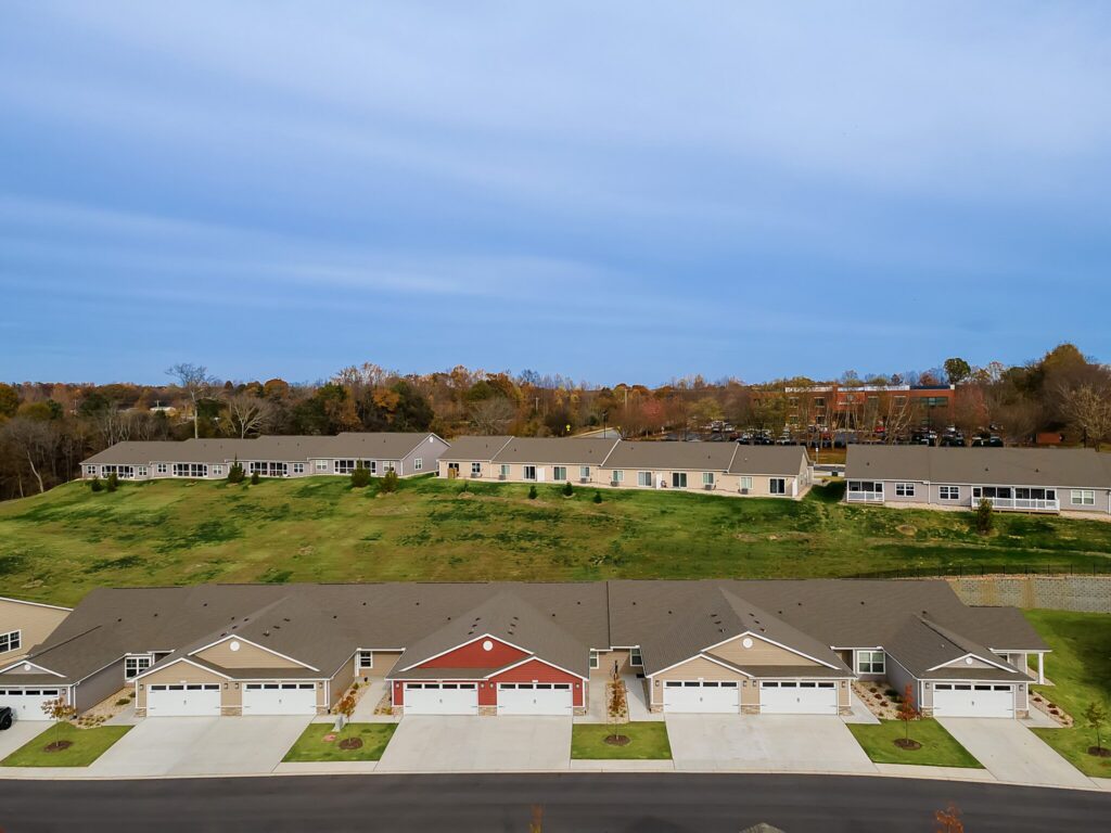 A row of suburban houses with driveways and garages is shown in the foreground, with grassy space and more buildings in the background under a clear blue sky.