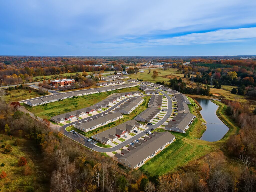 Aerial view of a suburban neighborhood with rows of houses, curving roads, a small pond, and surrounding trees with autumn foliage.