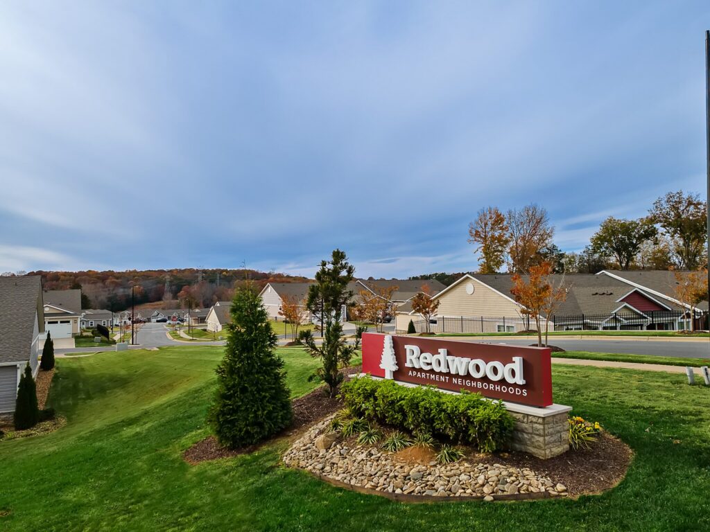 A red and white sign for Redwood Apartment Neighborhoods stands on a landscaped area beside a suburban street with houses in the background.