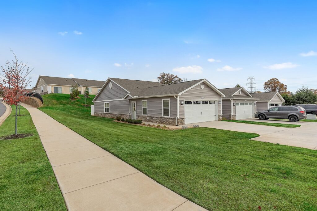 Single-story suburban houses with attached garages, well-maintained lawns, and a sidewalk under a clear blue sky. A car is parked in one driveway.