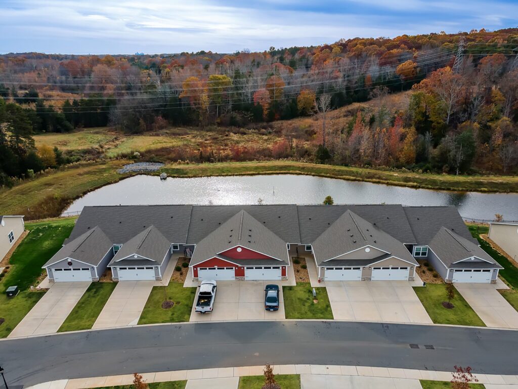 A row of attached suburban houses with driveways faces a small pond and wooded area in autumn, viewed from above.