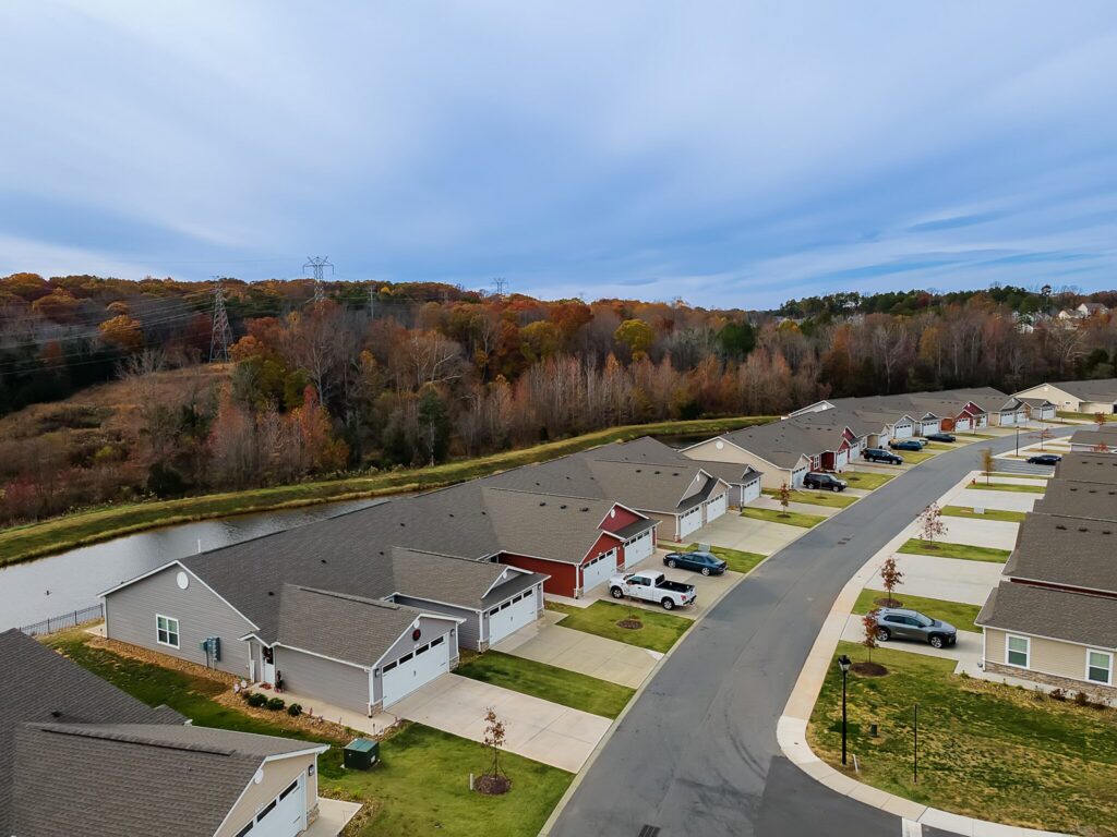 Aerial view of a suburban neighborhood with single-family homes, driveways, and cars parked along a curved street near a wooded area and a narrow waterway.