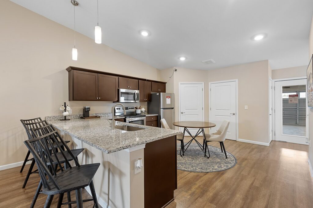 Modern kitchen with granite countertops, stainless steel appliances, brown cabinets, a small round dining table, and wooden flooring.