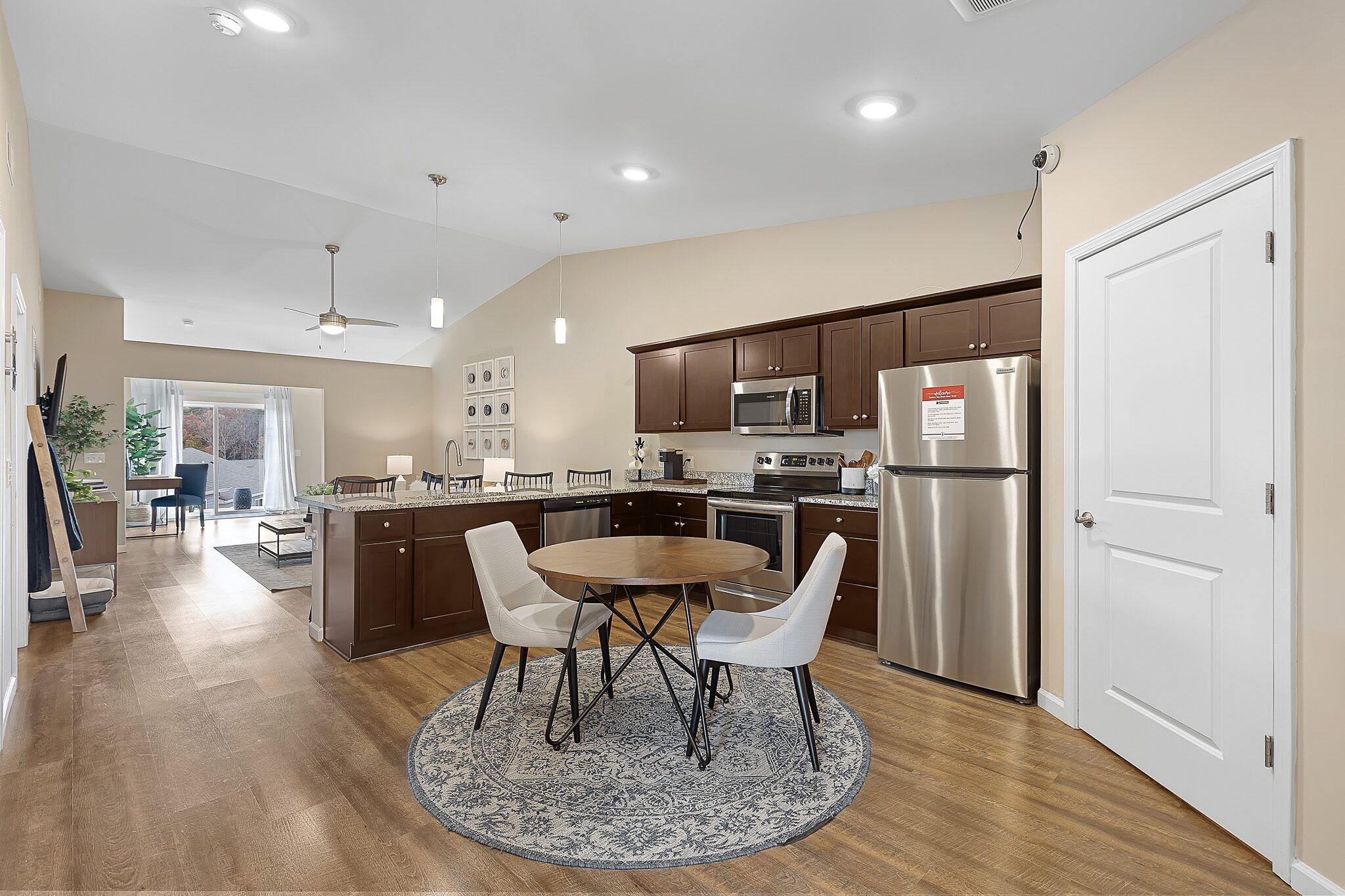 Modern open-concept kitchen and dining area with dark cabinets, stainless steel appliances, a round table with two chairs, and a living room visible in the background.
